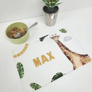 May include: A white acrylic placemat featuring a giraffe, green leaves, and gold flags. The name "MAX" is written in gold lettering.