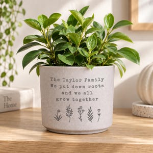 May include: A light grey cylindrical ceramic plant pot featuring the text The Taylor Family We put down roots and we all grow together in dark grey lettering, accompanied by five small botanical illustrations of various plants and flowers.