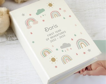 May include: Cream-colored photo album with a whimsical design featuring rainbows, clouds, suns, and stars. The album is personalized with the name "Doris" and the text "First Year of Adventures 2022-2023". The album is on a white wooden surface.