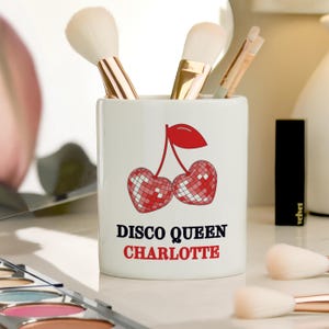 May include: A white ceramic makeup brush holder featuring a cherry design. The cherries are red and white with a disco ball pattern. The text "DISCO QUEEN CHARLOTTE" is printed below. Makeup brushes are inside the holder.