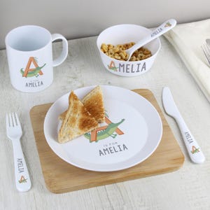 May include: A white children's breakfast set including a mug, bowl, plate, spoon, fork, and knife. Each item has a green alligator and the name "Amelia". Two pieces of toast sit on the plate.