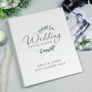 May include: A cream-coloured wedding photo album featuring the word "Wedding" in script, adorned with eucalyptus. The album is personalised with "Emily & James" and the date "17th August 2024". A camera and wedding photos are visible in the background.