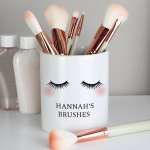 May include: White ceramic makeup brush holder with a black eyelashes design and the text "HANNAH'S BRUSHES". The holder is filled with various makeup brushes with rose gold handles.