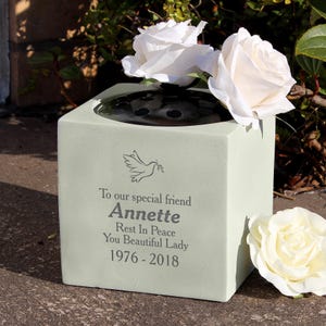 May include: A pale green square memorial vase with the name "Annette" and dates 1976-2018. The vase has a dove graphic and the words "To our special friend". White roses are placed in the vase.