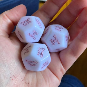 Puede incluir: Tres dados blancos de m&uacute;ltiples caras con texto rojo impreso en cada cara. El texto visible incluye frases como Tough Love, Kinesio Tape, Throw in Pool, Hugs, Hell if I know, Pelvic PT y Salon pas. Los dados tienen un acabado mate suave.