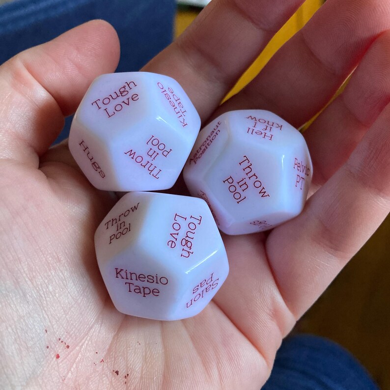 Puede incluir: Tres dados blancos de m&uacute;ltiples caras con texto rojo impreso en cada cara. El texto visible incluye frases como Tough Love, Kinesio Tape, Throw in Pool, Hugs, Hell if I know, Pelvic PT y Salon pas. Los dados tienen un acabado mate suave.