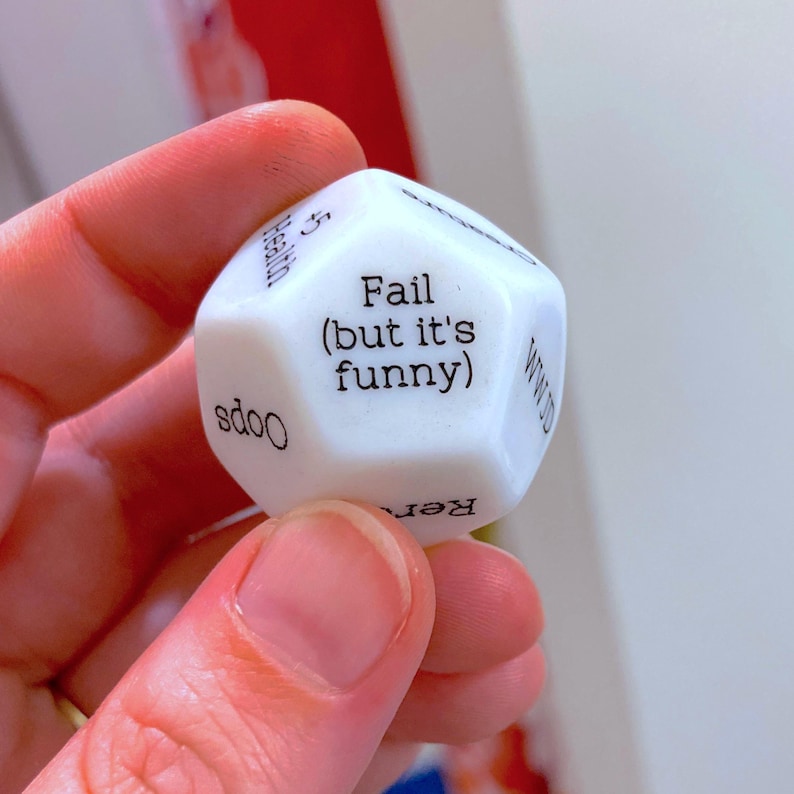 Puede incluir: Un dado blanco de doce caras con texto impreso en negro en cada cara. La cara central muestra el texto 'Fail (but it's funny)'. Otras caras visibles incluyen las palabras 'Health', 'Oops', 'Reroll' y 'WWJD' en una fuente clara sin remates.