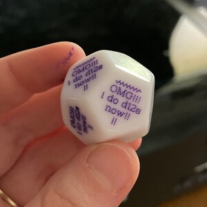 Puede incluir: Un dado de doce caras de color blanco con texto morado impreso en sus caras. El texto en los lados visibles dice "OMG!!! i do d12s now!! !!" con una l&iacute;nea decorativa de guiones sobre las letras. El dado tiene un acabado liso y brillante.