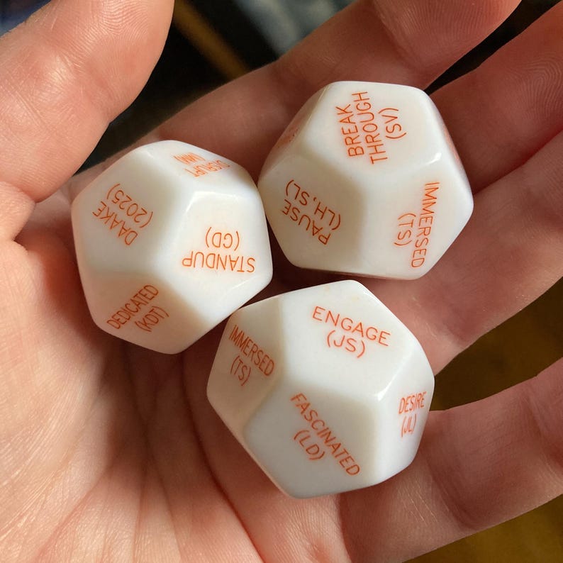 Puede incluir: Tres dados poli&eacute;dricos blancos con texto naranja impreso en cada cara. El texto incluye palabras como BREAK THROUGH, PAUSE, IMMERSED, STANDUP, DEDICATED, DAAKE 2025, ENGAGE, FASCINATED y DESIRE, cada una acompa&ntilde;ada de abreviaturas entre par&eacute;ntesis.
