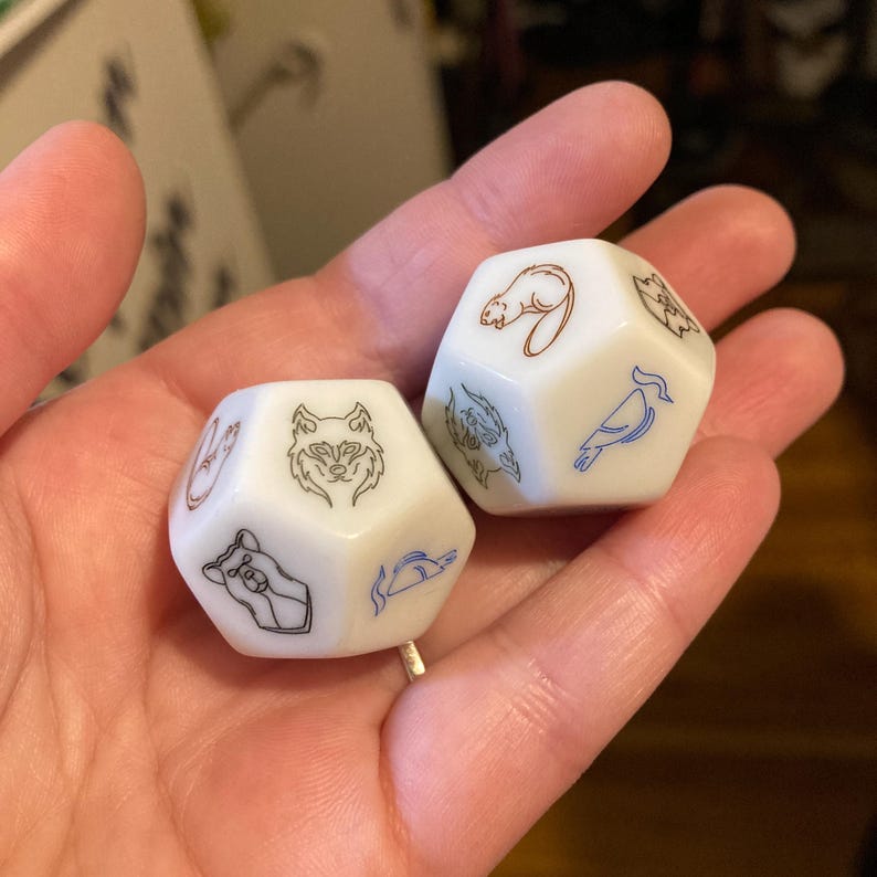 Puede incluir: Dos dados de juego blancos de varias caras con ilustraciones de animales grabadas. Un dado muestra una cabeza de lobo negra, una cabeza de oso y un pez azul. El otro dado muestra un castor marr&oacute;n, un p&aacute;jaro azul y una criatura negra.