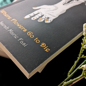 Może przedstawiać: Książka zatytułowana "Where Flowers Go to Die" autorstwa Sarah Marie Fasi. Okładka jest czarna z białą ilustracją dłoni i złotymi akcentami. Książka jest ułożona na innych książkach, z gałązką białych kwiat&oacute;w na pierwszym planie.