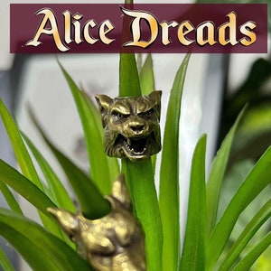 Könnte beinhalten: Zwei goldfarbene Tierkopf-Anhänger, einer von einem Wolf und einer von einem Hund, werden auf einer grünen Pflanze gezeigt. Die Pflanze steht in einem grauen Topf. Der Text "Alice Dreads" ist im Hintergrund sichtbar.