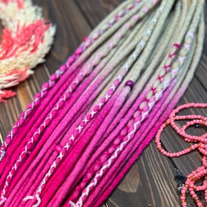 May include: A set of vibrant pink and beige dreadlocks, transitioning from a deep pink at the tips to a lighter beige at the roots. The dreads are adorned with white thread accents and small beads. A decorative wreath and beaded necklace are also visible.