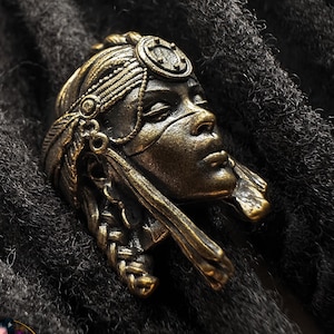 Peut inclure: Accessoire de dreadlocks de couleur bronze, représentant le profil détaillé d'un visage féminin orné d'une coiffe élaborée et d'éléments tressés. L'accessoire est mis en valeur sur un fond de dreadlocks gris foncé, créant un contraste saisissant.