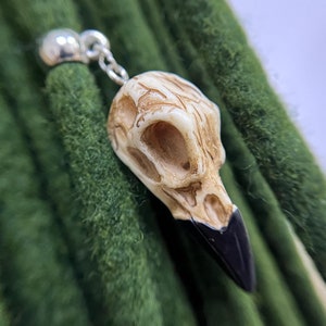 Puede incluir: Un amuleto de calavera de pájaro blanco con un pico negro, colgando de una cuenta y una cadena plateadas. El amuleto está sobre un fondo de rastas verdes.