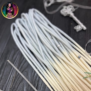 May include: A bundle of ombre synthetic dreadlocks with a light blue to white color gradient. The dreadlocks are arranged on a dark wooden surface with a small container of wax and two metal tools.