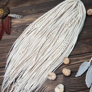May include: Off-white synthetic dreadlocks arranged on a dark wooden surface. The dreadlocks are long and have a natural appearance. Several seashells and decorative feathers are scattered around the dreadlocks. A logo with the text "Alice Dreads" is in the bottom right corner.