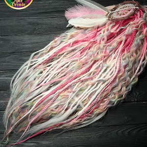 Op de afbeelding: Een verzameling kleurrijke dreadlocks in roze, witte en pastelkleuren. De dreadlocks zijn versierd met veren en een kleine dromenvanger. Het logo "Alice Dreads" is zichtbaar in de linkerbovenhoek.