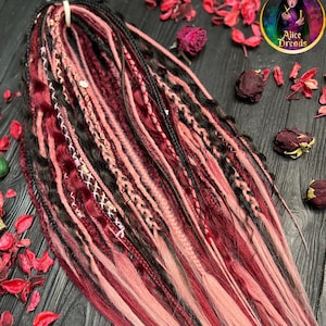 May include: A set of synthetic dreadlocks in shades of black, burgundy, and pink. The dreadlocks are arranged in a fan shape and are lying on a dark wooden surface.