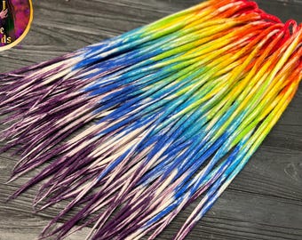 May include: Rainbow-colored dreadlocks, transitioning from red to yellow, green, blue, and purple. The synthetic hair extensions are arranged on a dark wooden surface. The Alice Dreads logo is visible in the upper left corner.