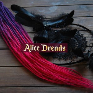 May include: A set of synthetic dreadlocks with a gradient color transition from dark purple to pink. The dreadlocks are displayed on a wooden surface with a dreamcatcher and feathers.