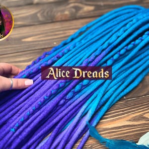 May include: A set of blue and purple ombre synthetic dreadlocks. The dreadlocks are made of wool and are approximately 24 inches long. The dreadlocks are arranged in a fan shape and are lying on a wooden surface.