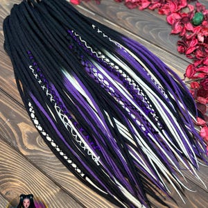 May include: A collection of black, purple, and white dreadlocks, featuring decorative stitching. The dreadlocks are displayed on a wooden surface. The colour scheme includes a gradient effect, transitioning from black to purple and white.