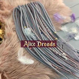 May include: A set of gray and light pink synthetic dreadlocks with white and brown accents. The dreadlocks are displayed on a pink fuzzy surface with feathers and beads.