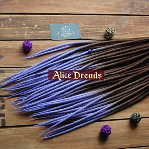 May include: A set of synthetic dreadlocks with a brown to purple ombre colour gradient. The dreadlocks are tied together with a blue ribbon and are displayed on a wooden surface.