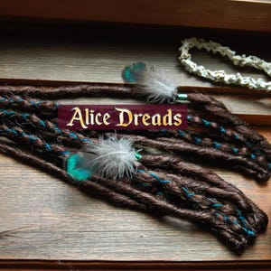 Peut inclure: Dreadlocks synthétiques marrons avec des accents turquoise et des plumes. Les dreadlocks sont présentées sur une surface en bois avec une pancarte qui indique "Alice Dreads".