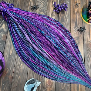 May include: A bundle of vibrant, multi-toned synthetic dreadlocks in shades of purple, teal, and pink, tied with a purple ribbon. The dreads are arranged on a wooden surface with decorative spiders and a logo that reads "Alice Dreads".