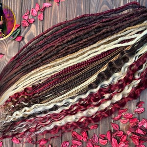 May include: A set of synthetic dreadlocks in various shades of brown, red, and blonde. The dreadlocks are arranged in a fan shape on a wooden surface with red flower petals scattered around.