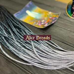 May include: A set of ombre synthetic dreadlocks in black and silver. The dreadlocks are made of kanekalon hair and are approximately 24 inches long. The dreadlocks are arranged in a fan shape on a wooden surface.