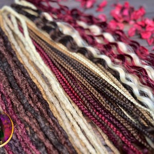 May include: A variety of synthetic dreadlocks in different colors and textures, including burgundy, brown, blonde, and black. The dreadlocks are arranged in a fan shape on a wooden surface.