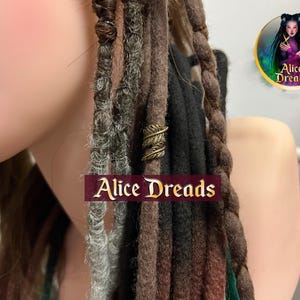 Peut inclure: Gros plan sur une variété de dreadlocks brunes, noires et grises avec un accent de perle en plume de métal doré. Les dreadlocks sont coiffées avec une variété de textures et d'épaisseurs. L'image comprend le texte "Alice Dreads".