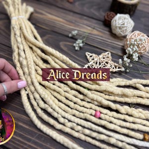 Puede incluir: Dreadlocks rubios, agrupados, con una cuenta rosa y una cuenta de madera. La imagen incluye el texto "Alice Dreads" en una pancarta granate. Elementos decorativos: estrellas y esferas tejidas.