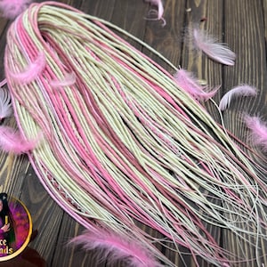 May include: A set of long, thin hair extensions featuring a blend of pale cream, soft pink, and vibrant magenta strands. The extensions are adorned with several light pink feathers and a small pink heart-shaped ornament with a lace-like pattern.