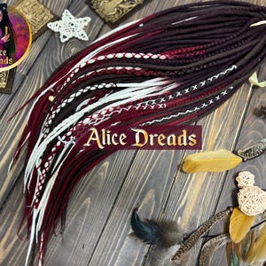 Op de afbeelding: Een set synthetische dreadlocks in de kleuren bordeaux, bruin en wit. De dreadlocks zijn in een losse vlecht gelegd en zijn met een wit lint aan elkaar gebonden. De dreadlocks liggen op een houten oppervlak.