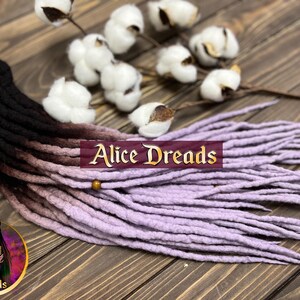 May include: A set of long, ombre dreadlocks with black, brown, and lavender colors. The dreadlocks are made of wool and are arranged in a fan shape. The dreadlocks are on a wooden surface with cotton balls in the background. The text "Alice Dreads" is visible in the image.