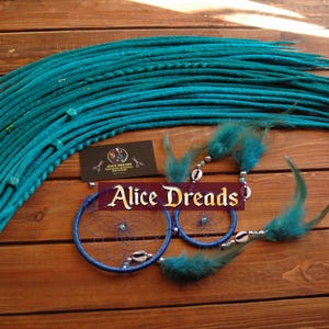 May include: Teal blue synthetic dreadlocks with a blue and white dreamcatcher and feathers. The dreamcatcher has the text "Alice Dreads" on it.