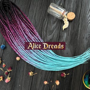 May include: A set of synthetic dreadlocks with a gradient color scheme, transitioning from dark purple to light blue. The dreadlocks are displayed on a dark wooden surface.