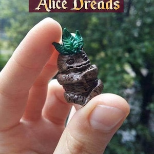 Könnte beinhalten: Eine kleine, handbemalte Figur aus braunem Ton, die eine Kreatur mit grünen Blättern auf dem Kopf darstellt. Die Kreatur hat einen runden, braunen Körper mit einem Gesicht und Armen. Die Figur wird in der Hand einer Person gehalten.