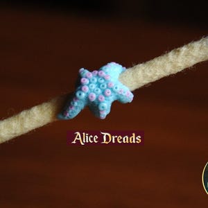 Puede incluir: Una cuenta de estrella de mar azul claro con detalles rosas enhebrada en un dreadlock de hilo beige y azul. El texto "Alice Dreads" es visible debajo de la cuenta.