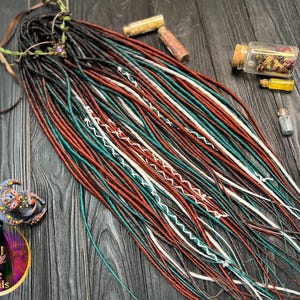 May include: A set of synthetic dreadlocks in shades of brown, teal, white, and red. The dreadlocks are arranged in a fan shape on a wooden surface.
