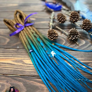May include: A bundle of handmade dreadlocks in shades of brown, green, and vibrant blue, tied with a purple ribbon. The dreads are adorned with beads and charms. The background includes pine cones and feathers on a wooden surface. The logo "Alice Dreads" is visible.
