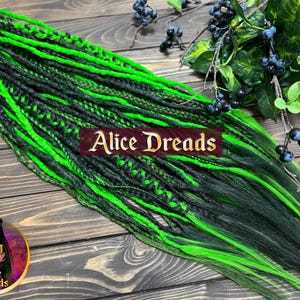 Puede incluir: Dreadlocks negros y verde neón con el texto "Alice Dreads". Las rastas están dispuestas sobre una superficie de madera con follaje decorativo y bayas. Un logotipo circular con una mujer y el texto "Alice Dreads" está en la esquina inferior izquierda.