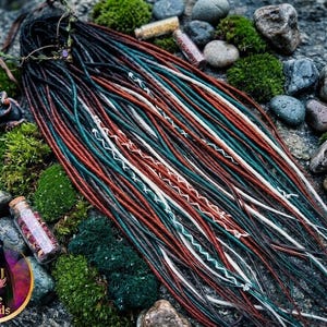 Puede incluir: Un conjunto de rastas sintéticas con una paleta de colores negro, verde bosque profundo y rojo óxido. Los mechones están acentuados con hilos blancos enrollados y cordones decorativos. Un pequeño frasco de vidrio con pétalos secos se encuentra cerca.