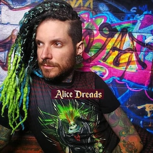 May include: A person with green and blue dreadlocks is wearing a black t-shirt with a graphic print of a skull with green and black accents. The text "Alice Dreads" is visible on the shirt. The person is standing in front of a graffiti wall.