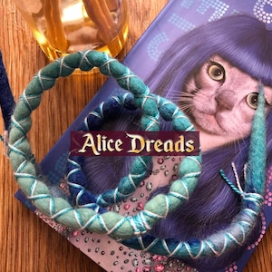 May include: Two blue and green wool dreadlocks with white accents. The dreadlocks are on a book with a purple haired cat on the cover. The book has the text "Alice Dreads" on it.