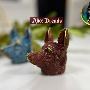 May include: A close-up of a red, gold and blue ceramic animal head sculpture. The sculpture is of a wolf or dog with a pointed snout and erect ears. The sculpture is on a white surface.